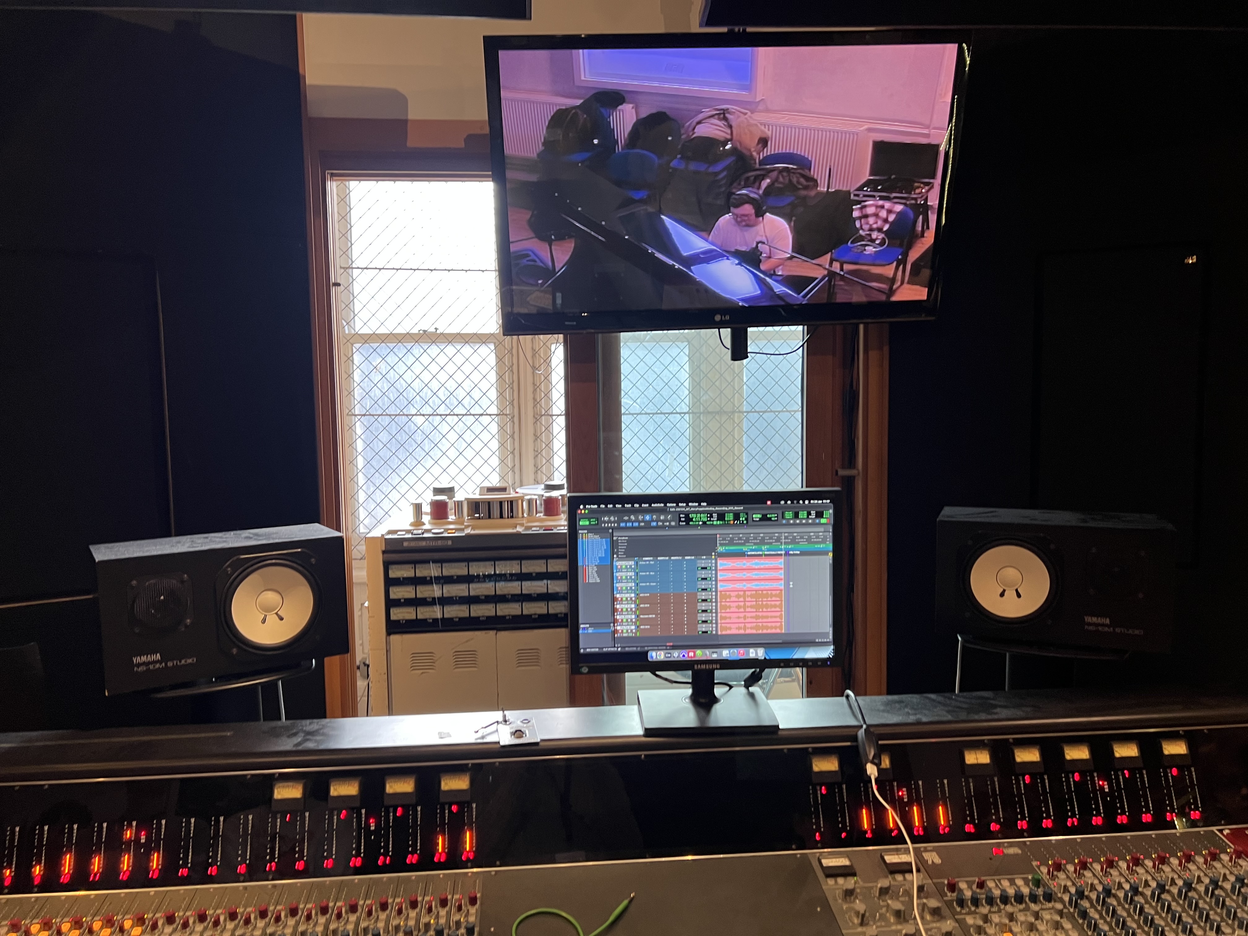 A Neve Custom 75 mixing console, NS10 speakers and Pro Tools running in the background. A TV screen mounted above the console shows a video feed of a pianist.